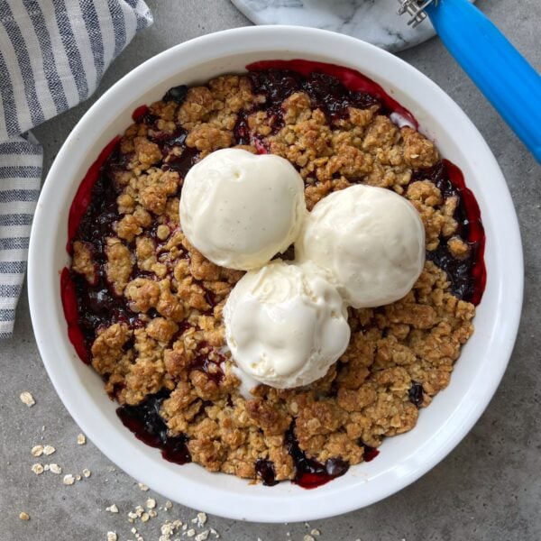 A round dish of berry crumble topped with three scoops of vanilla ice cream sits on a gray surface next to a striped cloth.