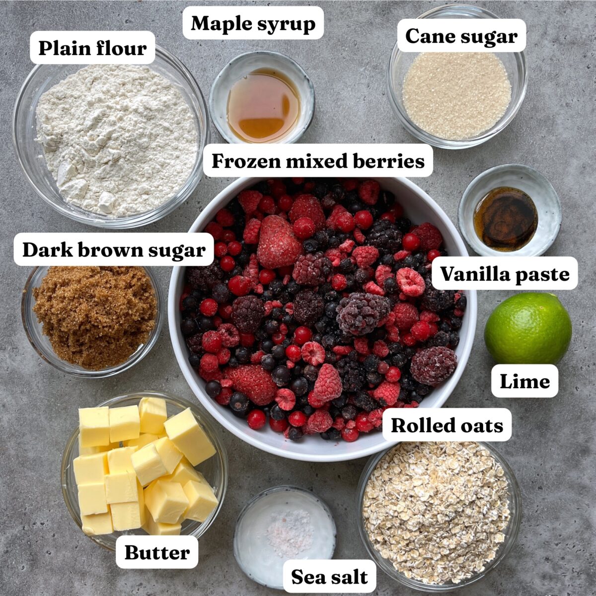 A bowl of frozen mixed berries is surrounded by ingredients including flour, maple syrup, cane sugar, dark brown sugar, vanilla paste, lime, rolled oats, butter, and sea salt on a gray surface.