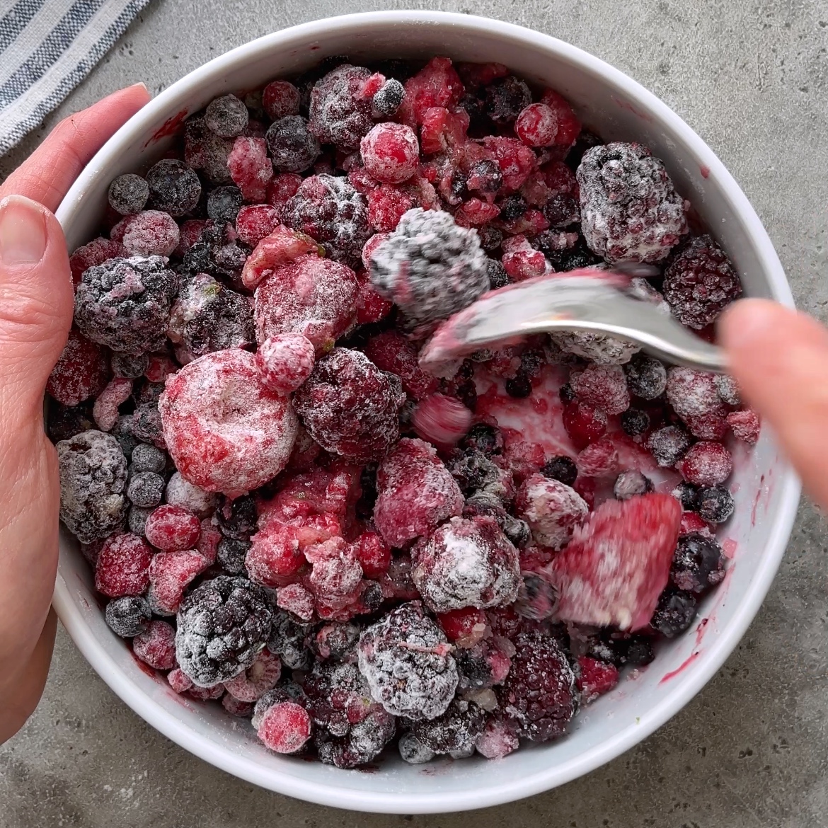 A bowl of frozen mixed berries being stirred with a spoon on a gray surface.