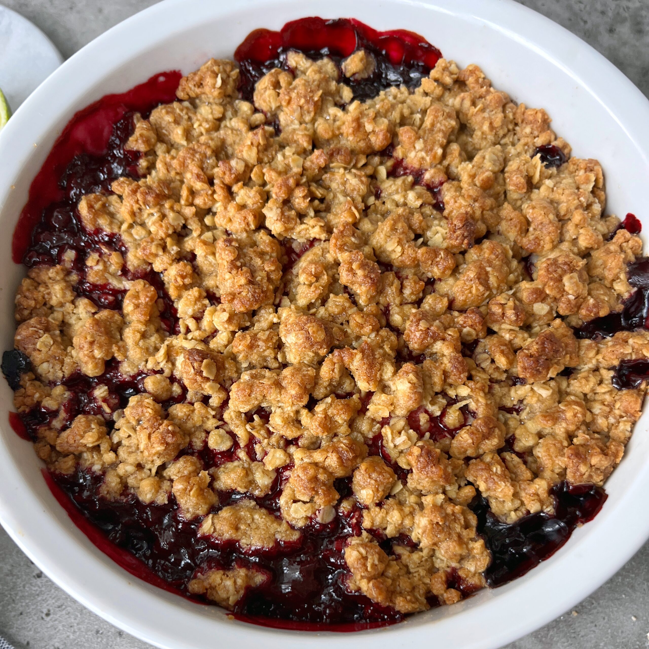 A baked mixed berry crumble with a golden brown oat topping and bubbling berry filling in a round white dish.