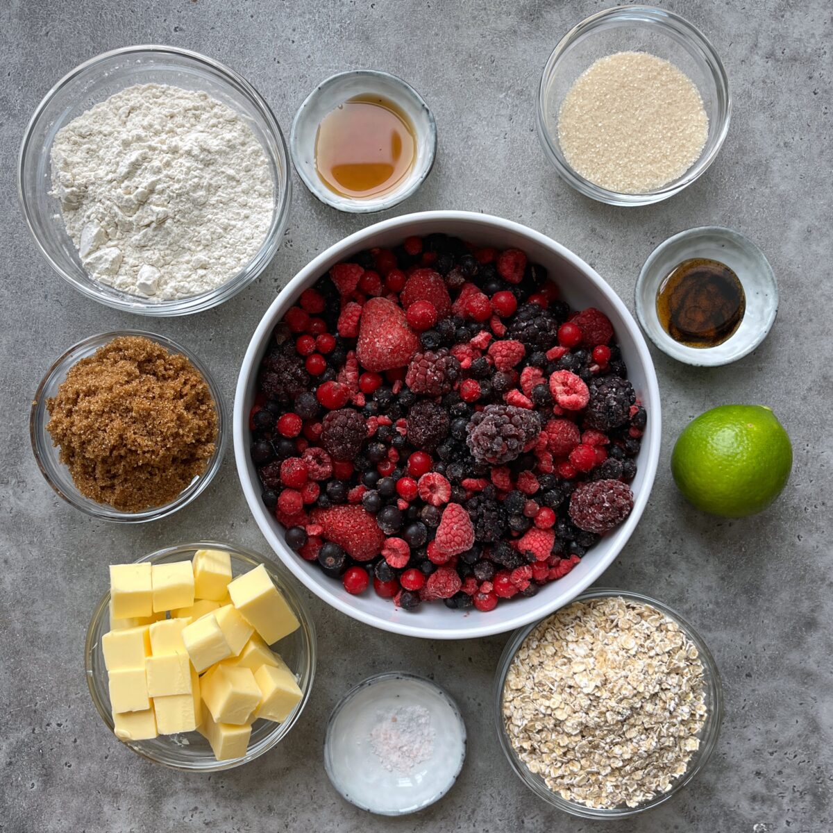 Ingredients for a berry crumble recipe arranged on a gray surface, including oats, sugars, butter, and syrup in glass bowls, with frozen mixed berries, flour, lime, and vanilla paste.