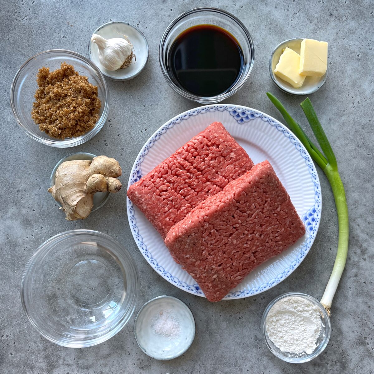 Ingredients for a recipe arranged on a gray surface, including ground beef, garlic, ginger, brown sugar, soy sauce, butter, green onion, flour, salt, and a small bowl of water.