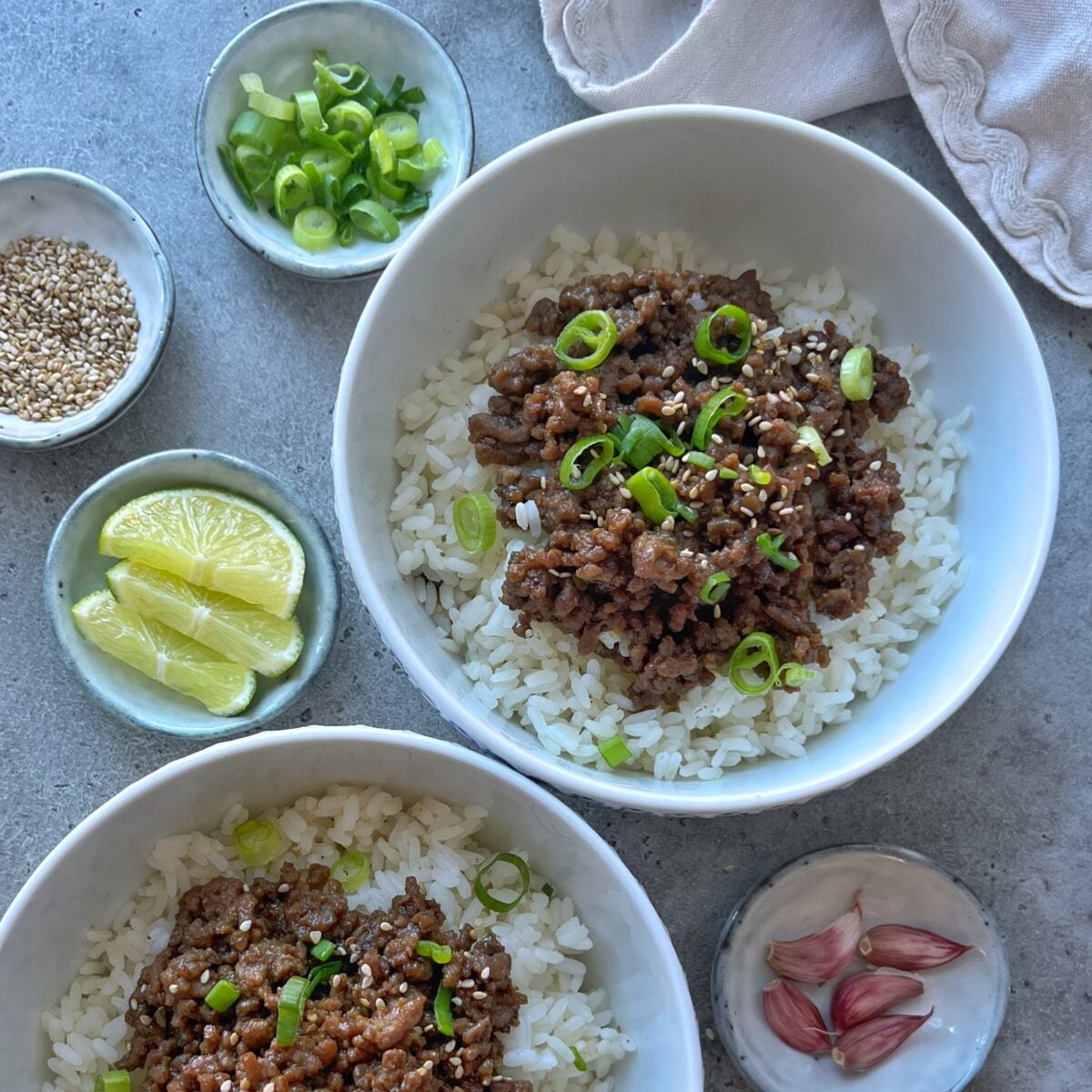 Two bowls of white rice topped with savory Mongolian Ground Beef and chopped green onions, surrounded by small dishes of lime wedges, sesame seeds, green onions, and garlic cloves.