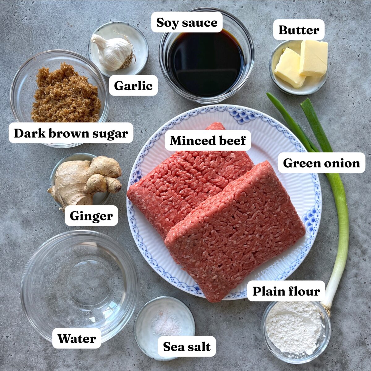 Top-down view of ingredients including minced beef, soy sauce, butter, dark brown sugar, garlic, green onion, ginger, plain flour, water, and sea salt on a gray surface.