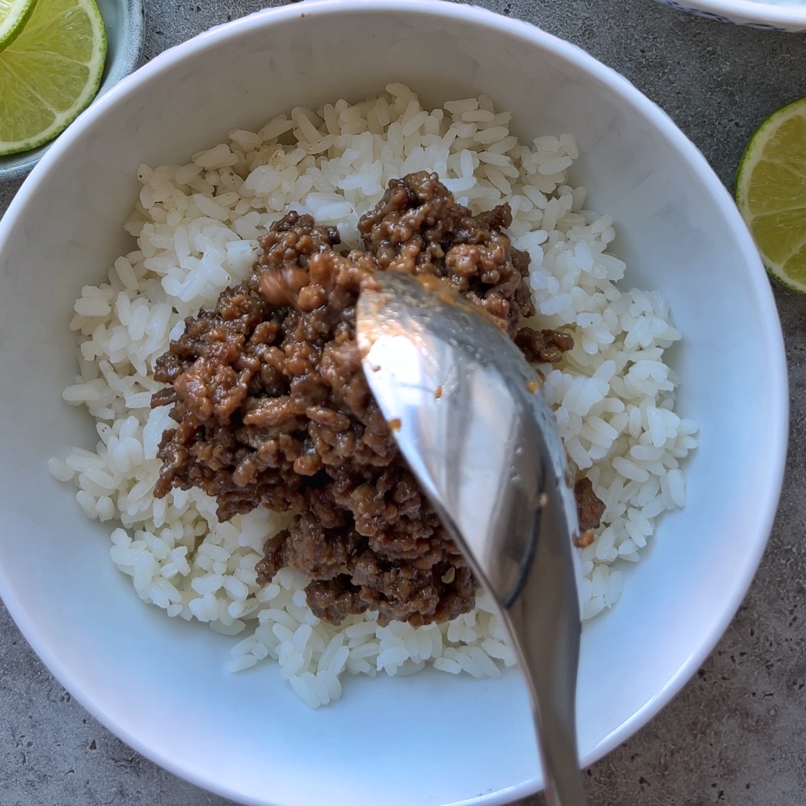 A spoon is serving Mongolian Ground Beef onto a bowl of white rice, with lime slices visible on the side.