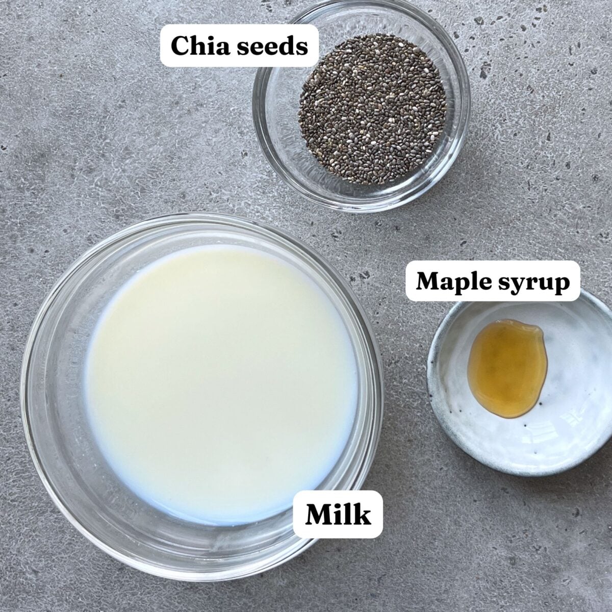 Three labeled bowls on a gray surface display the key ingredients for Overnight Chia Pudding: chia seeds, milk, and maple syrup.