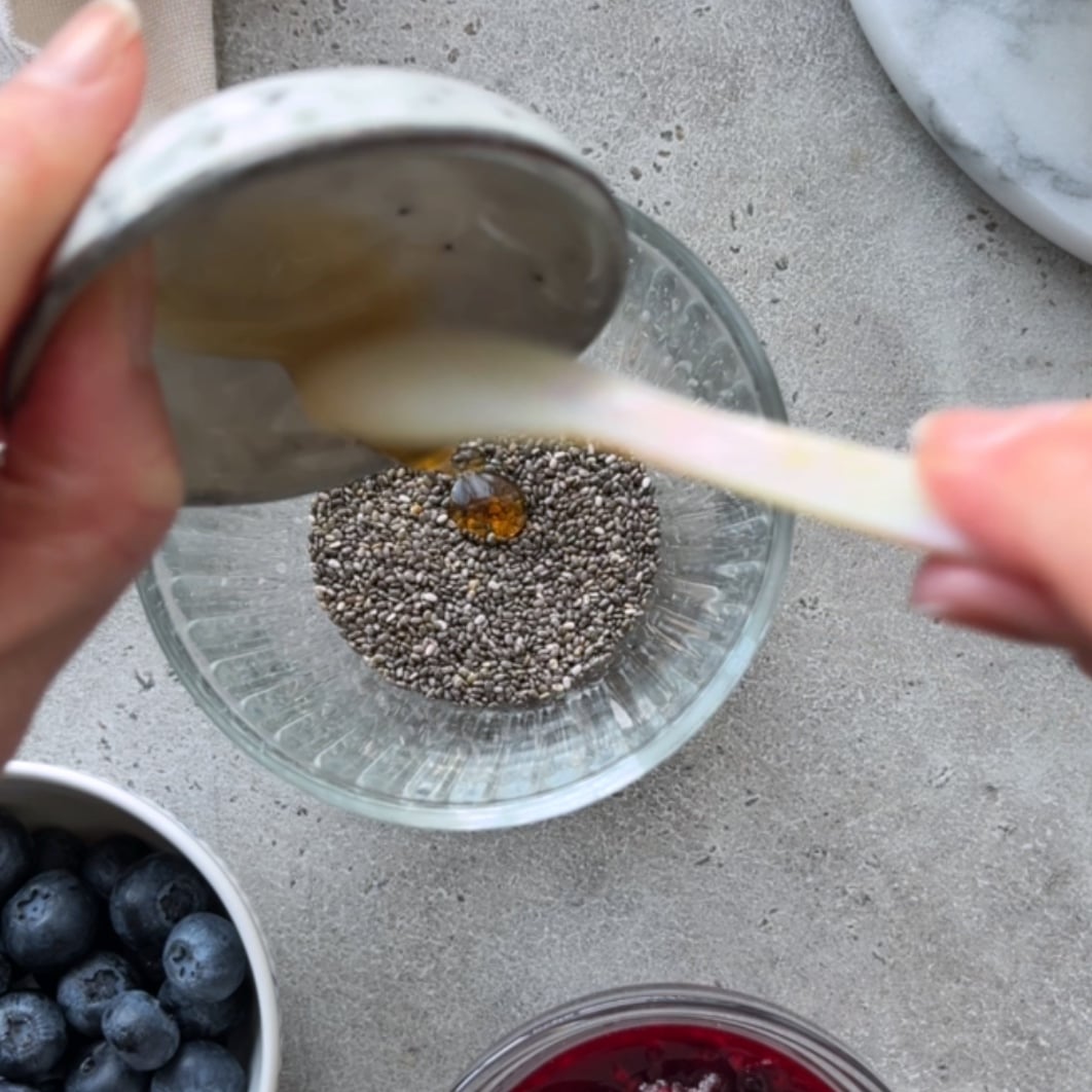 A person pours maple syrup into a glass filled with chia seeds, preparing Overnight Chia Pudding, while bowls of blueberries sit nearby on a gray countertop.