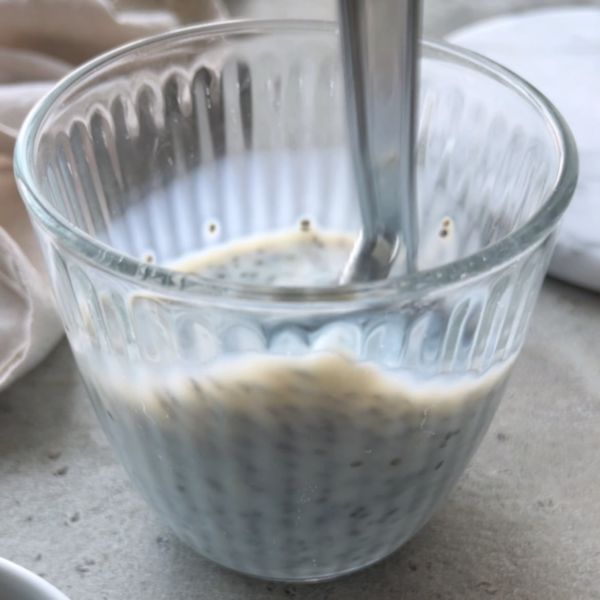 A spoon stirs a glass of chia seeds soaked in a milk, placed on a light-colored surface.