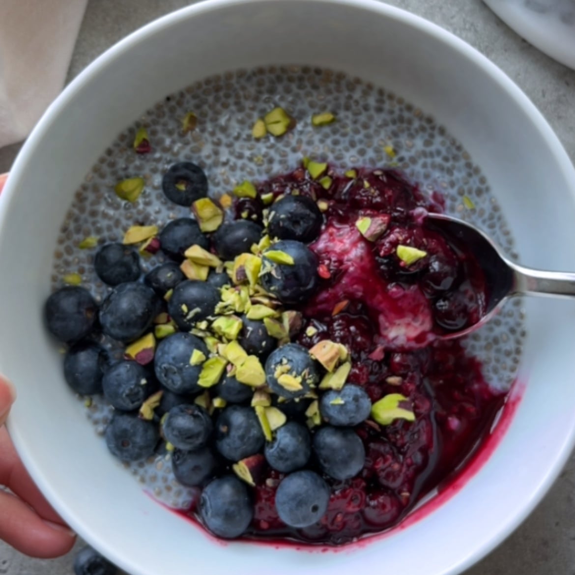 A spoon digging into a bowl of Overnight Chia Pudding topped with blueberries, berry compote, Greek yogurt, and chopped pistachios.