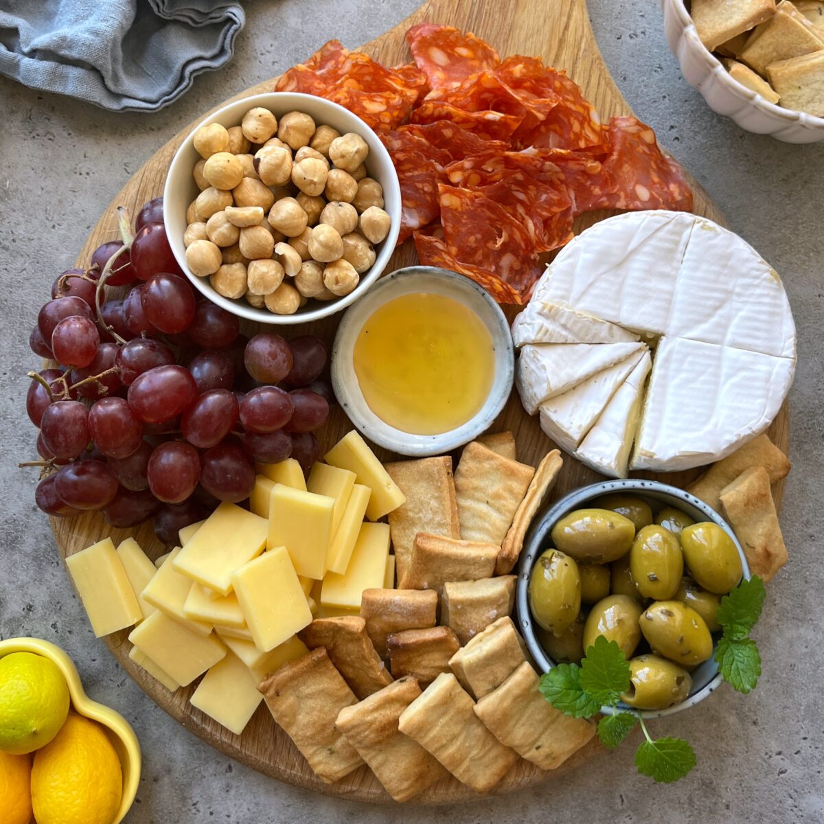 A wooden board with sliced cheese, crackers, green olives, red grapes, hazelnuts, sliced cured meat, a wheel of brie cheese, and a small dish of honey.