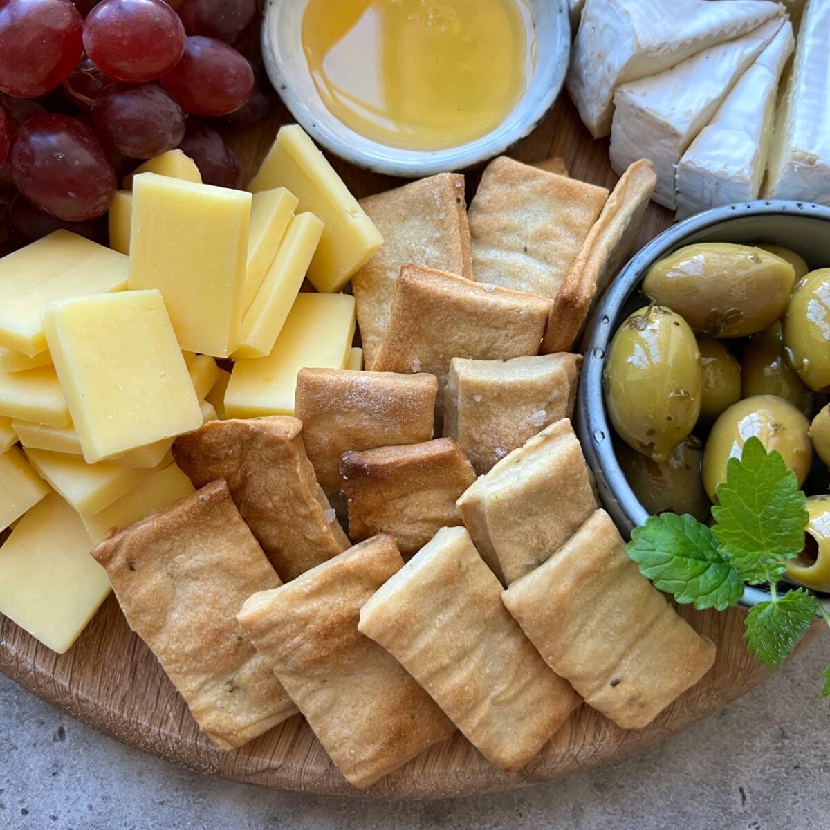 A wooden board with sliced cheese, grapes, honey, crackers, green olives in a bowl, and wedges of brie cheese.