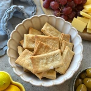 A bowl of rectangular crackers on a table, next to grapes, cheese cubes, lemons, and green olives.