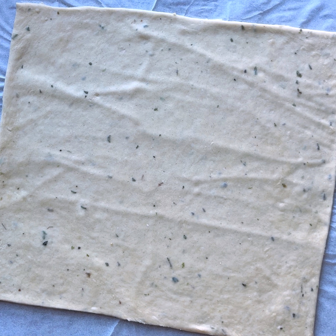 A square sheet of uncooked dough with visible herbs.