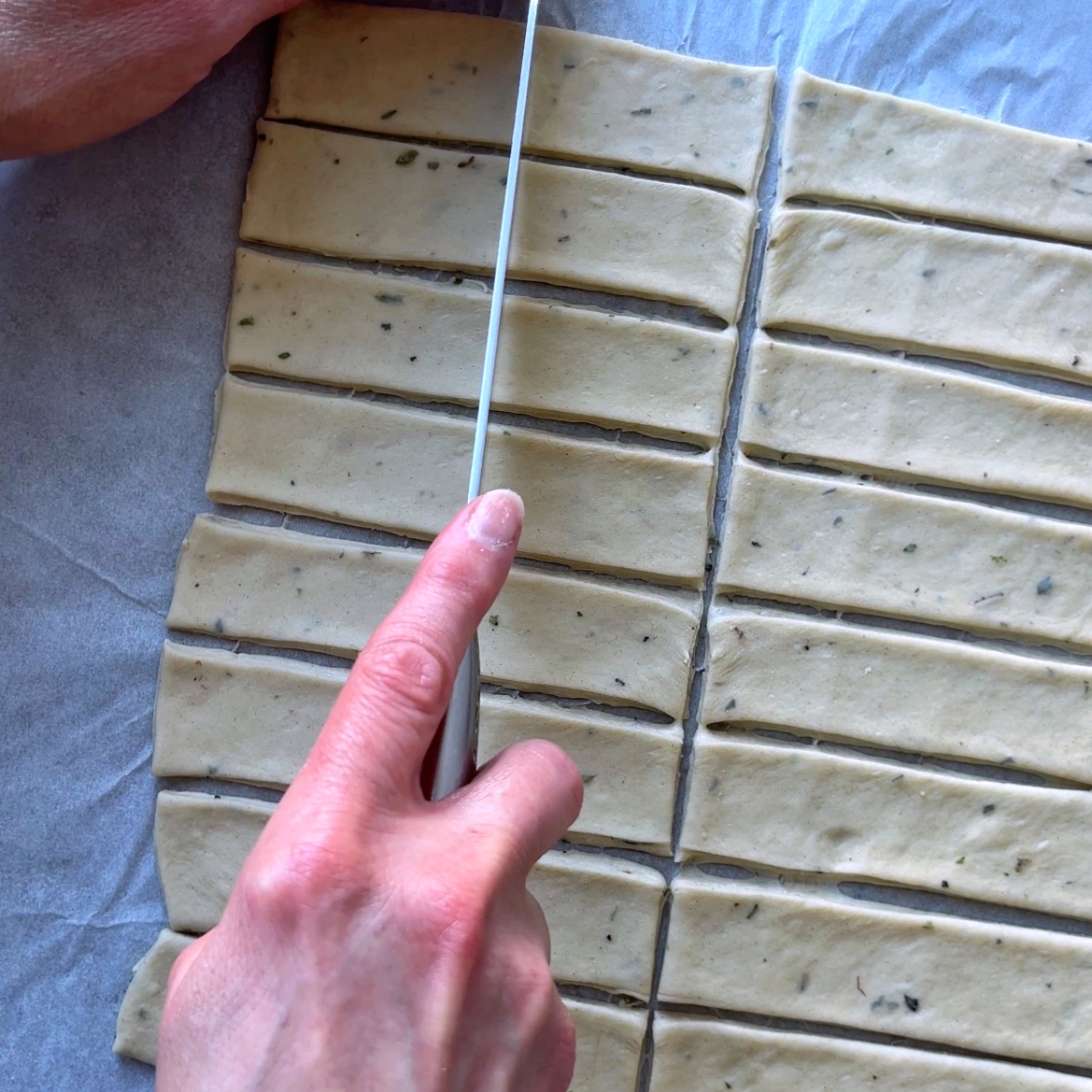 A person uses a knife to cut herbed dough into evenly sized rectangular strips on parchment paper.