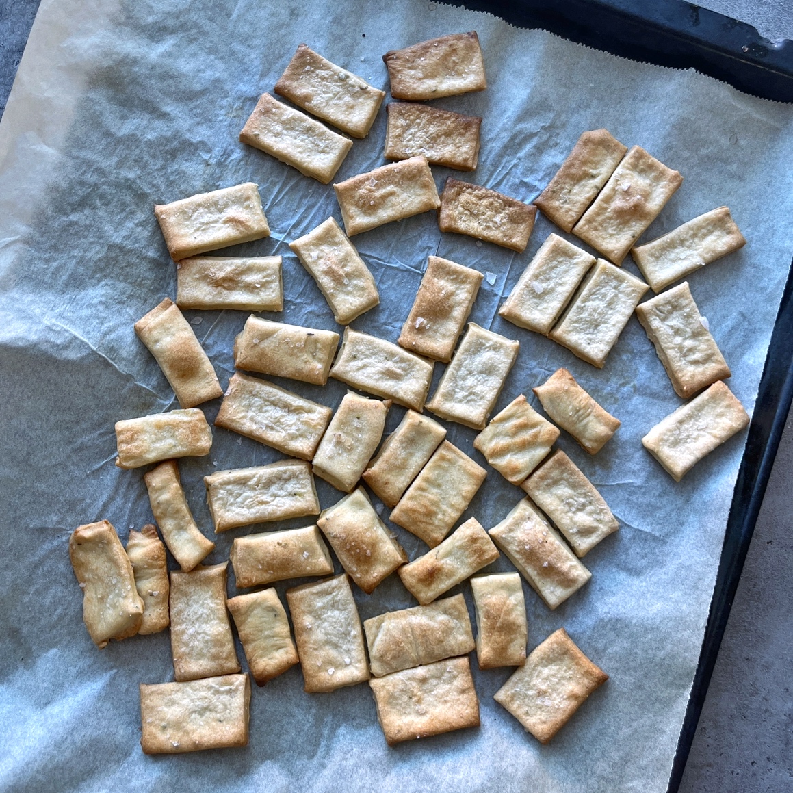 Artisan Crackers, rectangular and homemade, are spread out on a parchment-lined baking tray, some overlapping slightly.