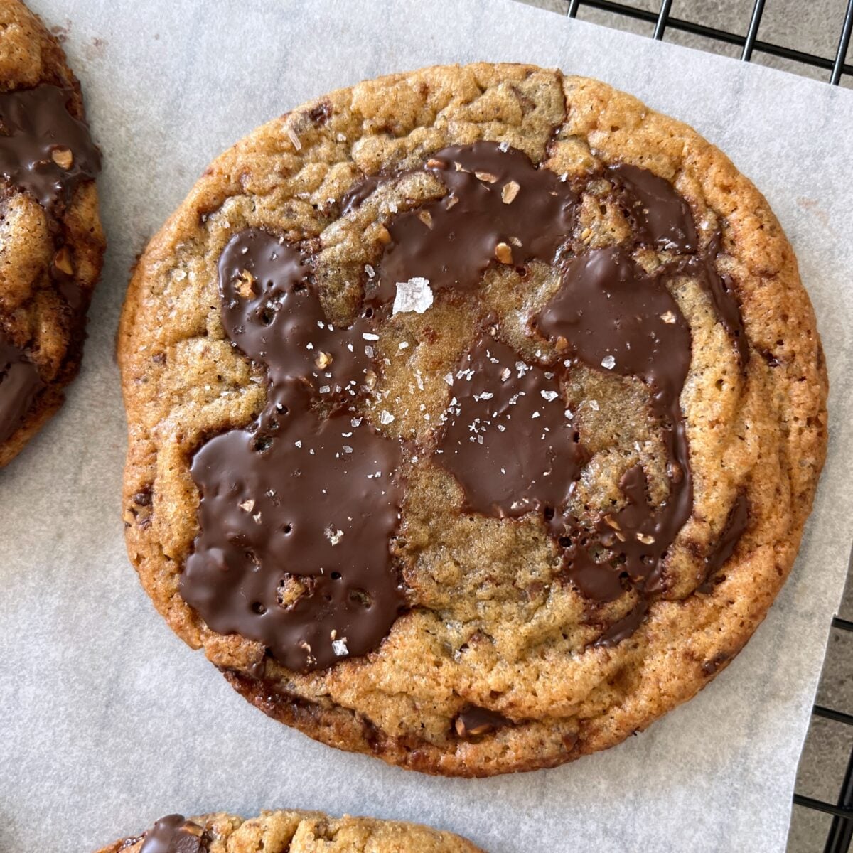 A large Best Chewy Chocolate Chip Cookie with melted chocolate patches and a sprinkle of flaky sea salt sits on parchment paper.