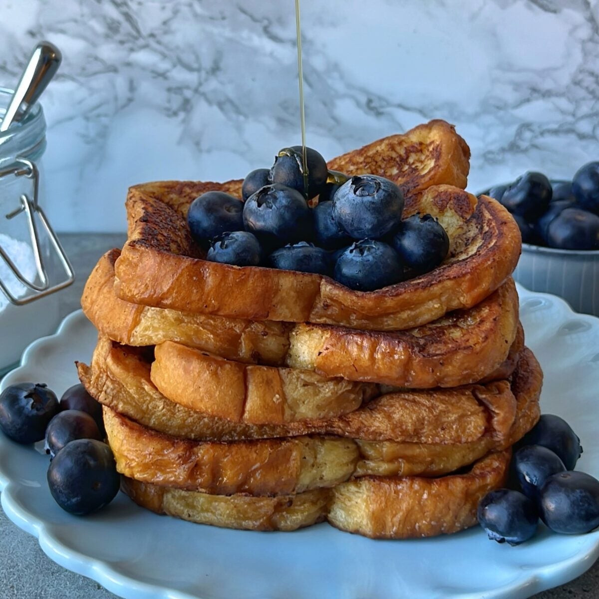 A stack of Brioche French Toast topped with fresh blueberries, with syrup being poured over, served on a white plate.