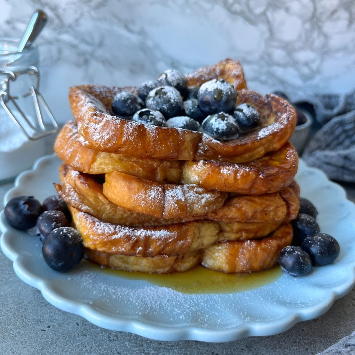 A stack of Brioche French Toast slices topped with blueberries and powdered sugar on a white plate, with syrup and extra blueberries around the base.