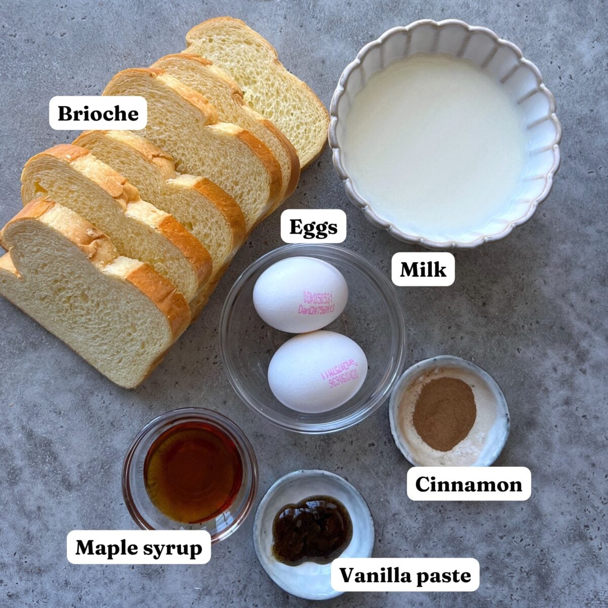 Brioche French Toast ingredients: Sliced brioche, two eggs, a bowl of milk, cinnamon, maple syrup, and vanilla paste arranged on a gray surface with labels.