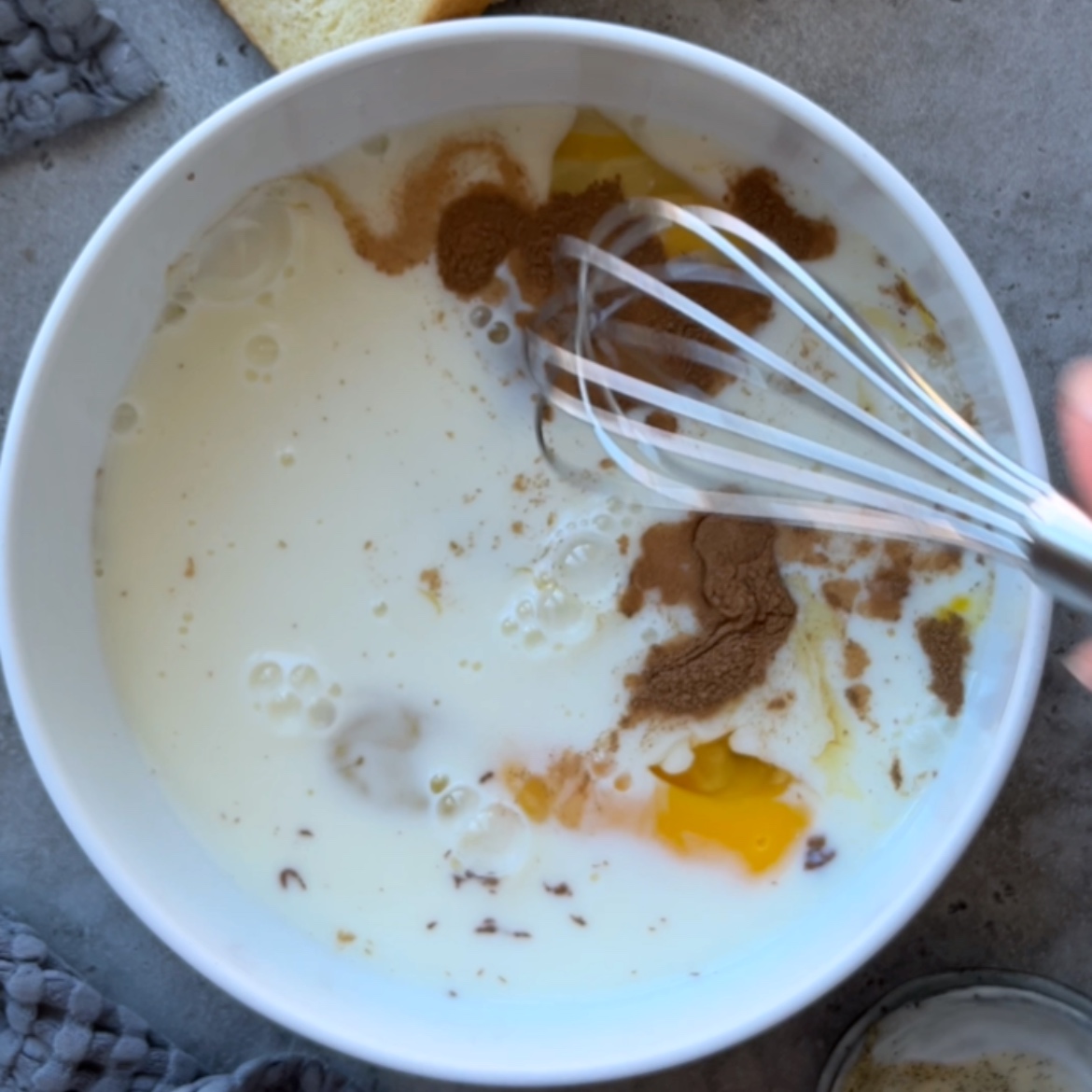 A whisk stirring eggs, milk, and spices in a white bowl, preparing a delicious mixture for Brioche French Toast.