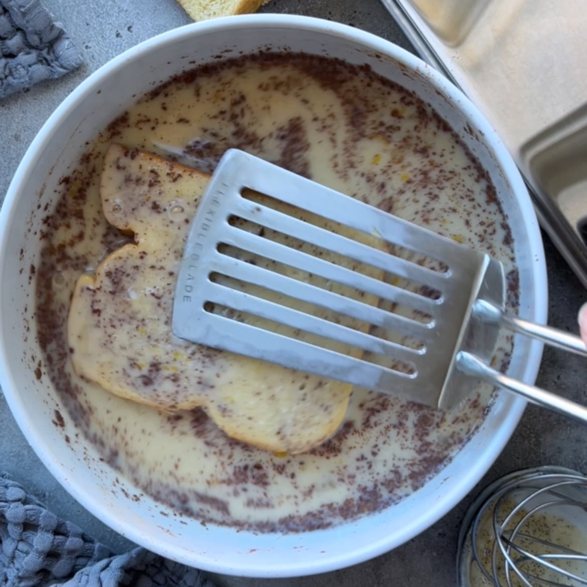 A metal spatula pushes sliced brioche into a white dish filled with an egg and milk mixture.