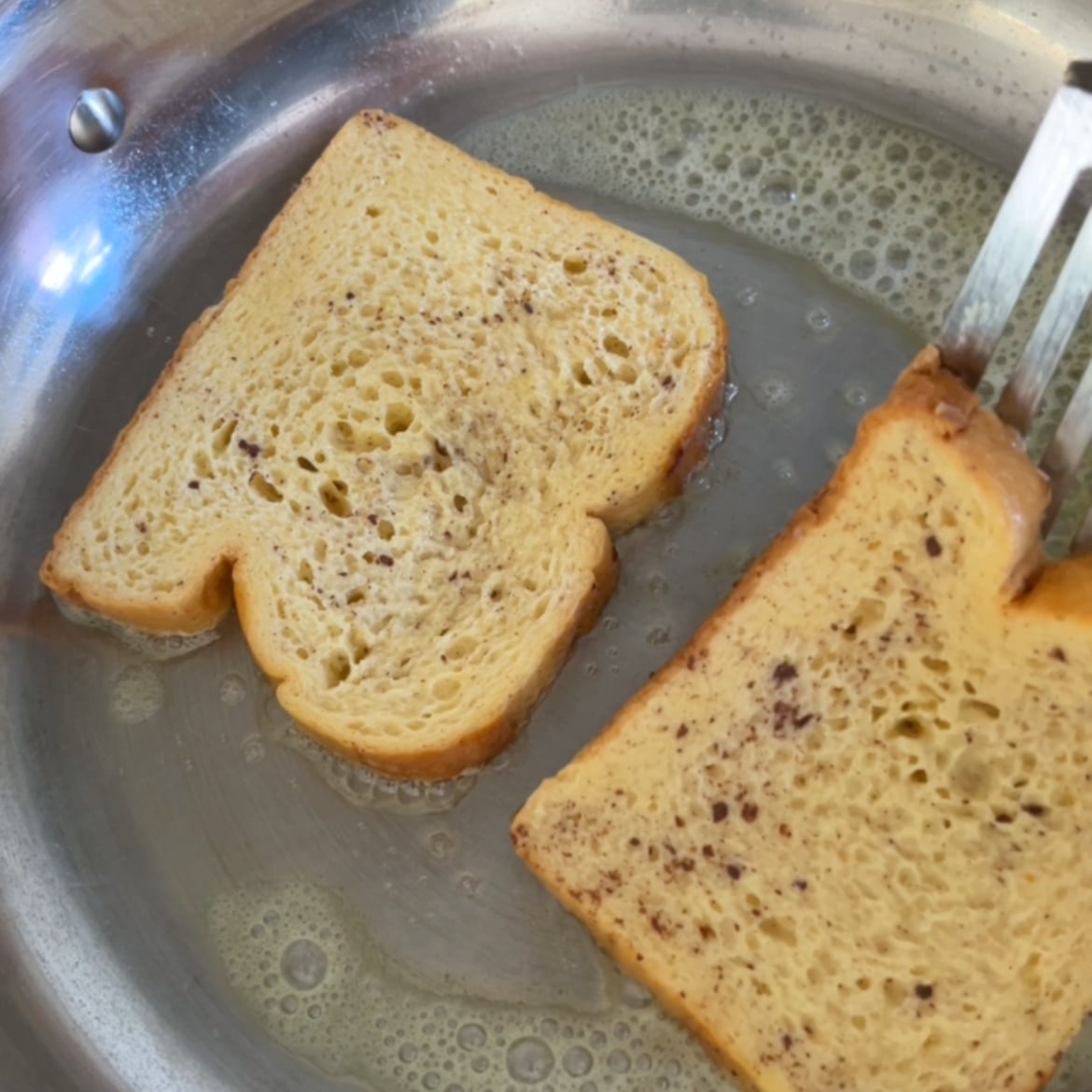 Two slices of Brioche French Toast coated in egg mixture are frying in a stainless steel pan, with a spatula lifting one  slice.