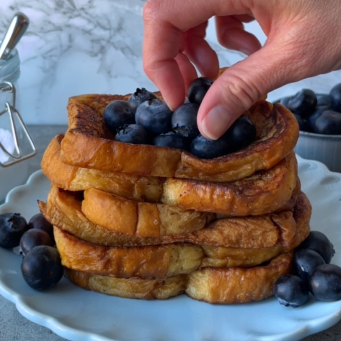 A hand places fresh blueberries atop a stack of Brioche French Toast slices on a light blue plate, with more blueberries scattered around.