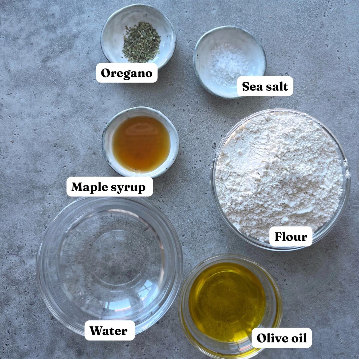 Six labeled bowls on a gray surface contain oregano, sea salt, maple syrup, flour, water, and olive oil.