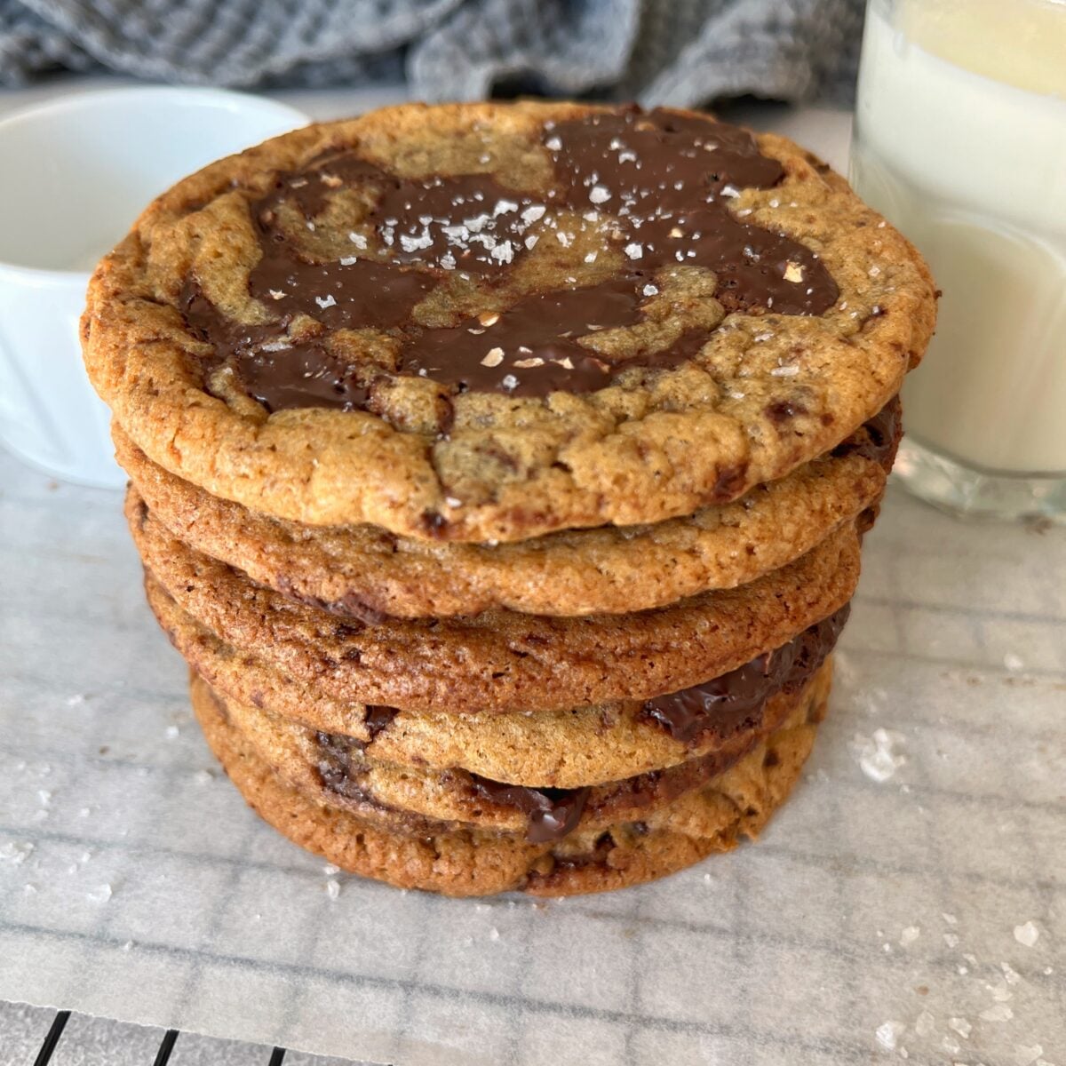 A stack of six chewy chocolate chip cookies topped with sea salt sits on parchment paper next to a glass of milk.