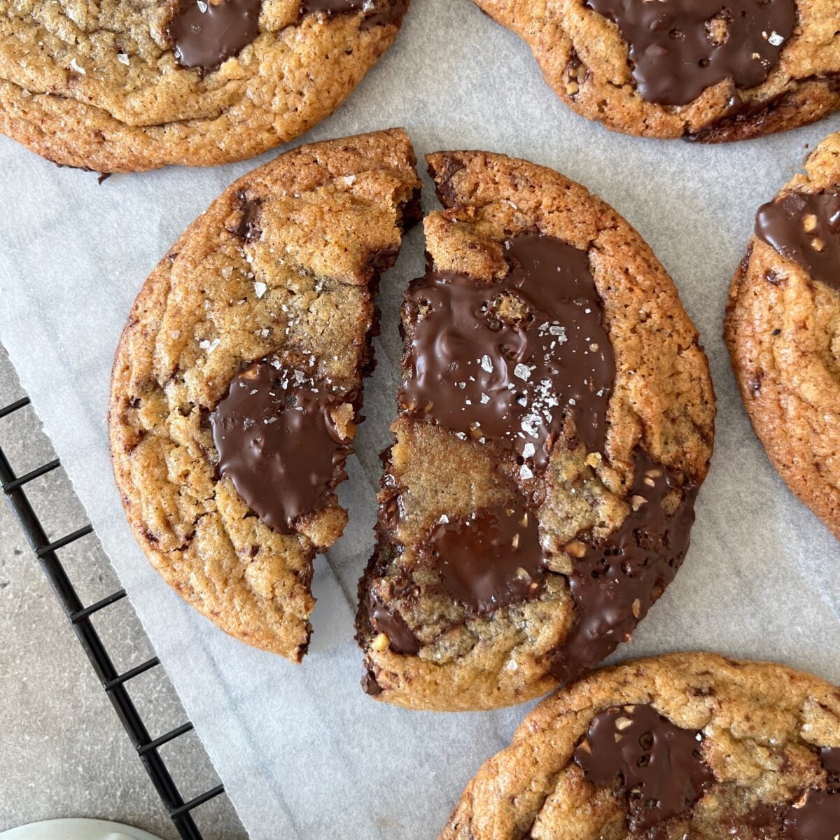 A chewy chocolate chip cookie, broken in half, rests on parchment paper with visible pools of melted chocolate and a light sprinkle of sea salt on top.