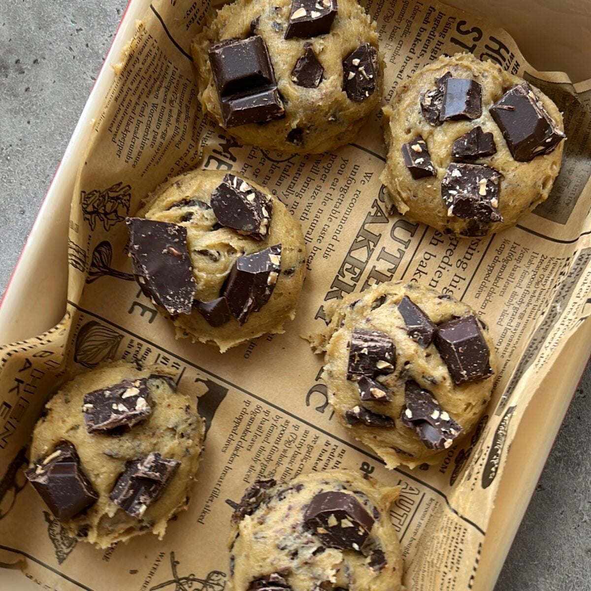 Six unbaked Chewy Chocolate Chip Cookies with chocolate chunks rest on parchment paper, arranged in a rectangular baking dish.