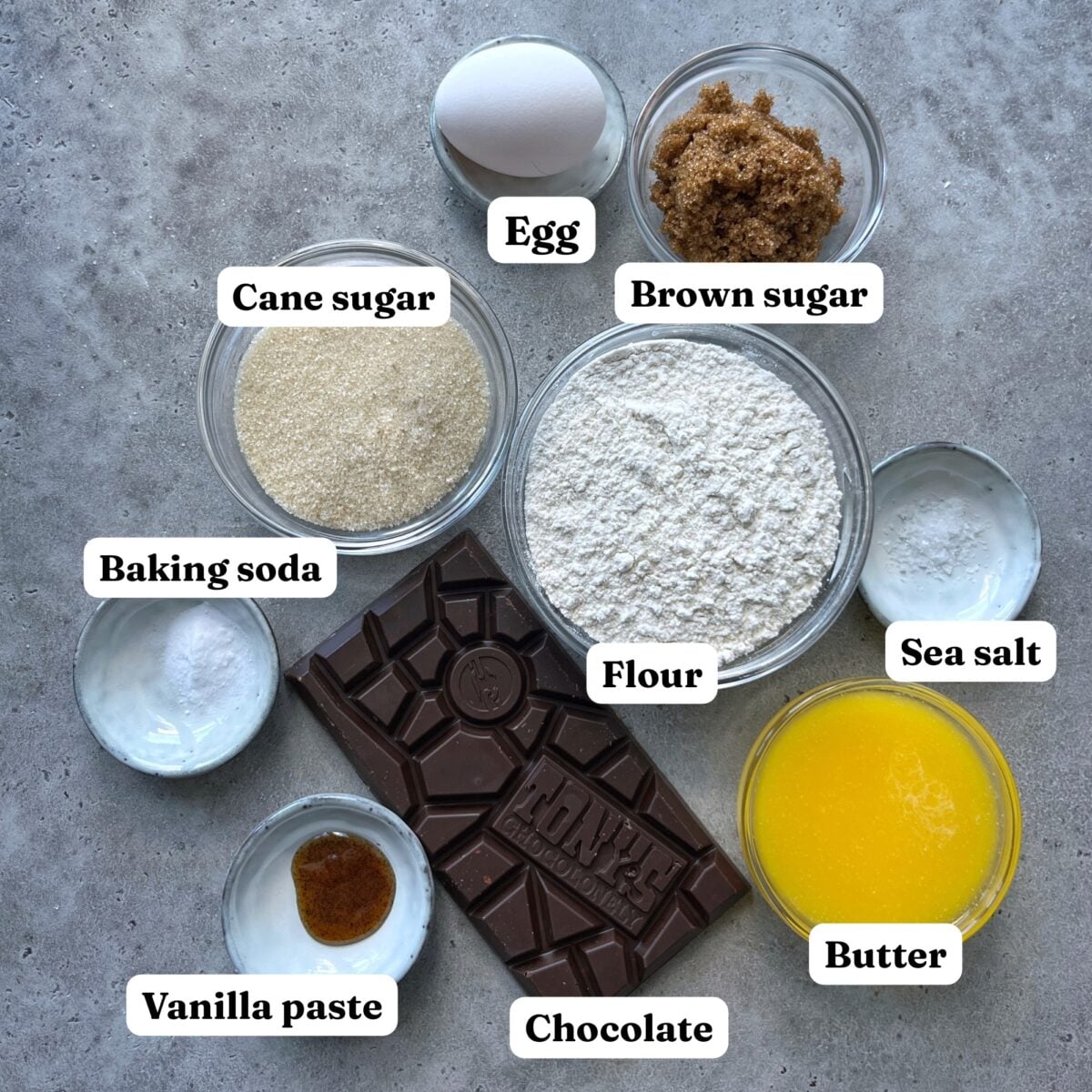 Ingredients for baking Chewy Chocolate Chip Cookies: an egg, cane sugar, brown sugar, baking soda, flour, sea salt, vanilla paste, a chocolate bar, and melted butter are arranged on a gray surface.