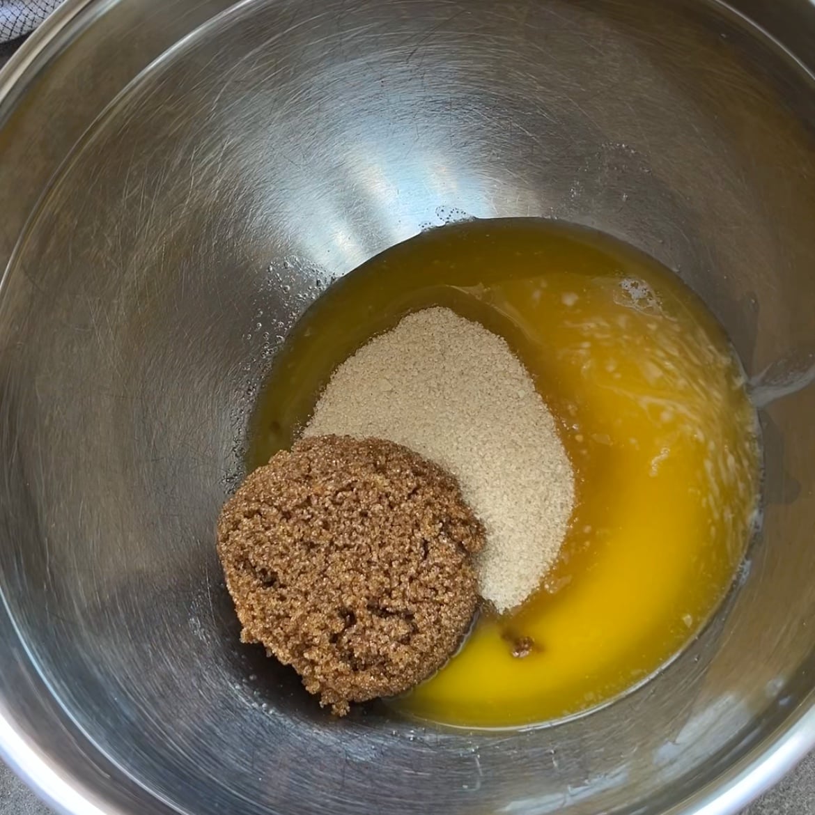 A metal mixing bowl containing melted butter, brown sugar, and cane sugar.