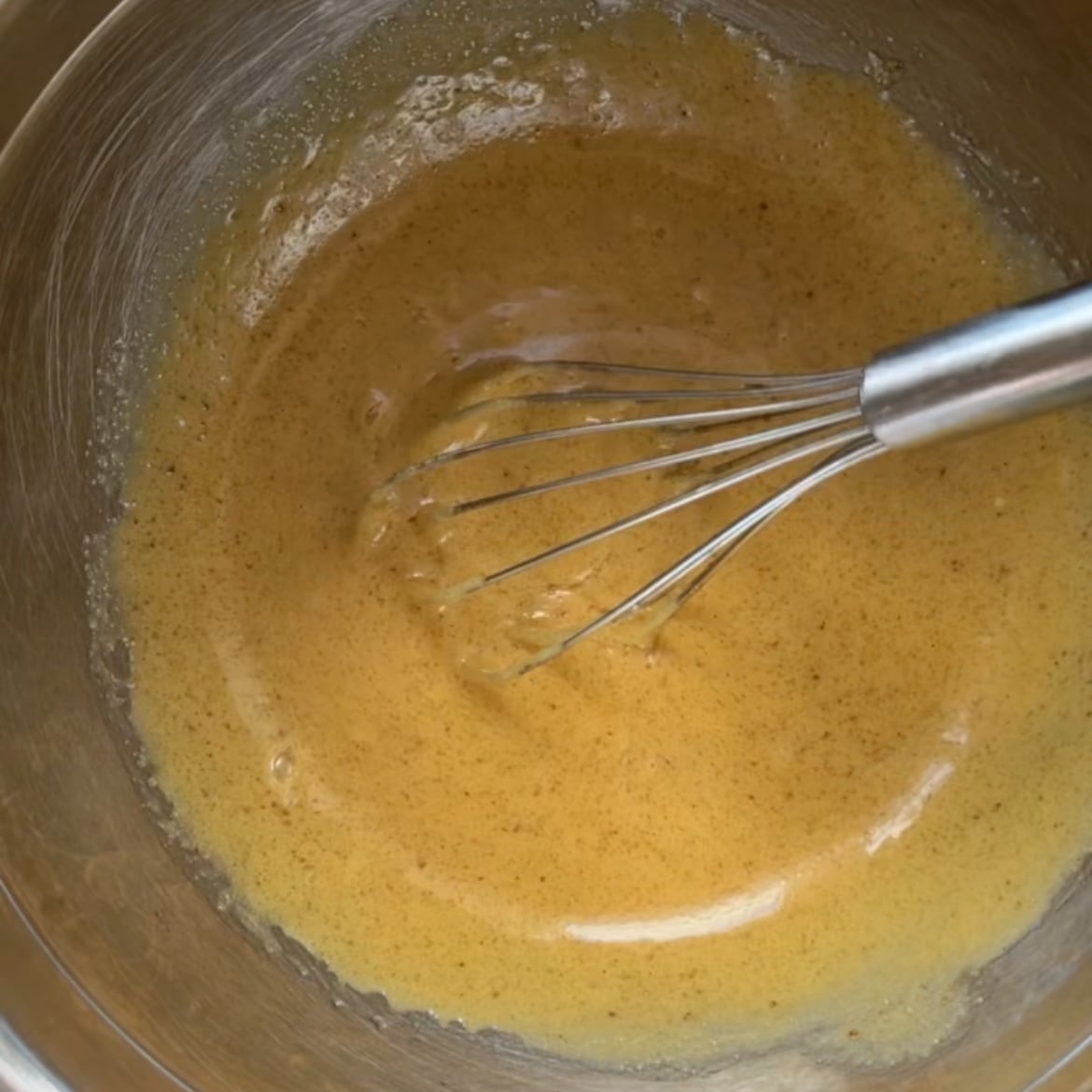 A metal whisk mixing a light brown batter with eggs, vanilla, melted butter, and two types of sugar in a stainless steel bowl.