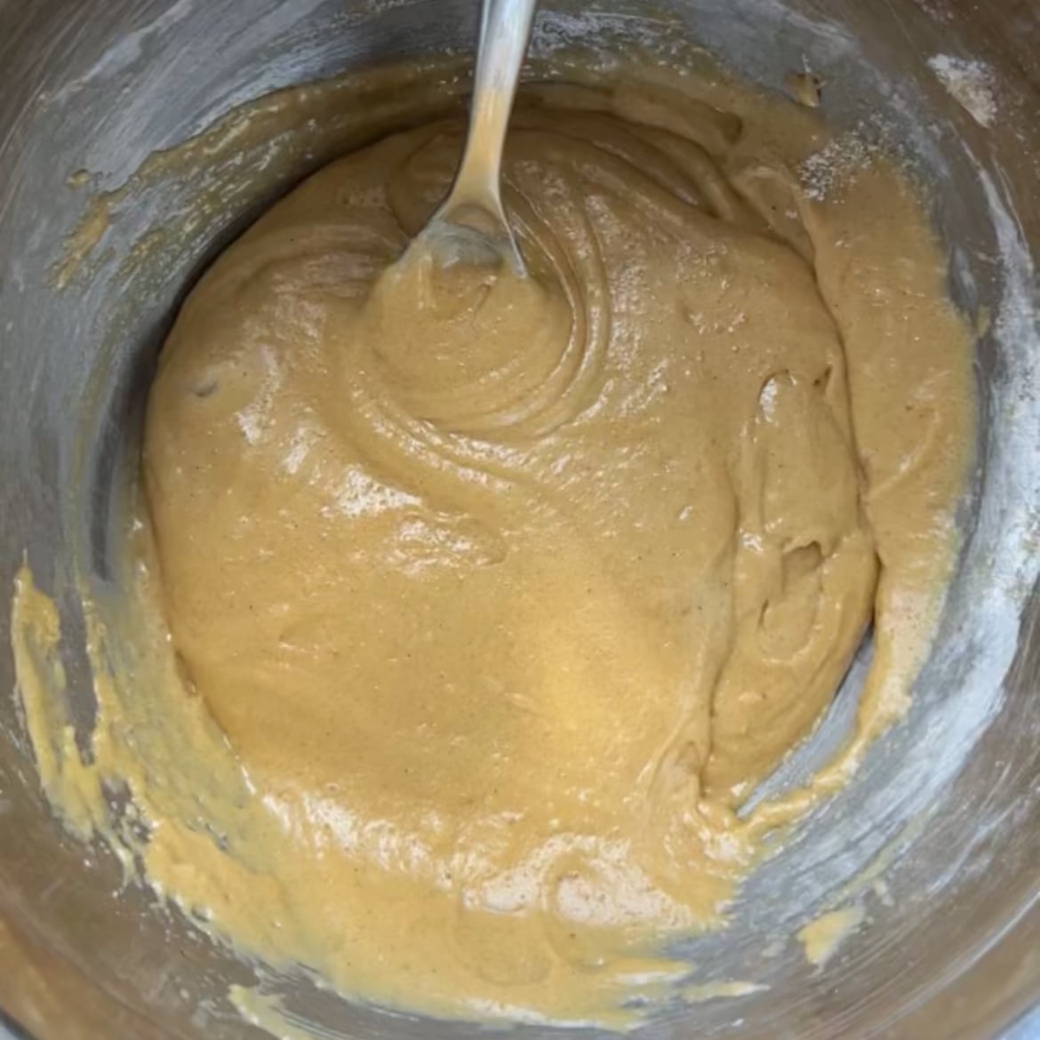 A metal bowl filled with thick, light brown batter being mixed with a spoon.