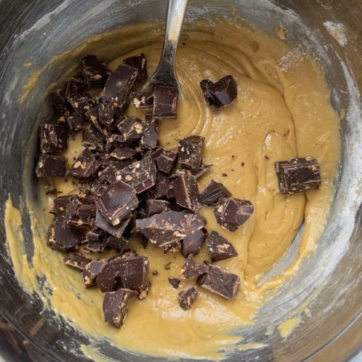 A metal bowl with cookie dough being mixed by a spoon, topped with chunks of dark chocolate.