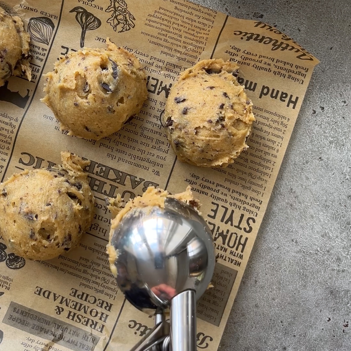 Scoops of chewy chocolate chip cookie dough sit on a sheet of printed parchment paper, with a metal cookie scooper holding another portion in the foreground.