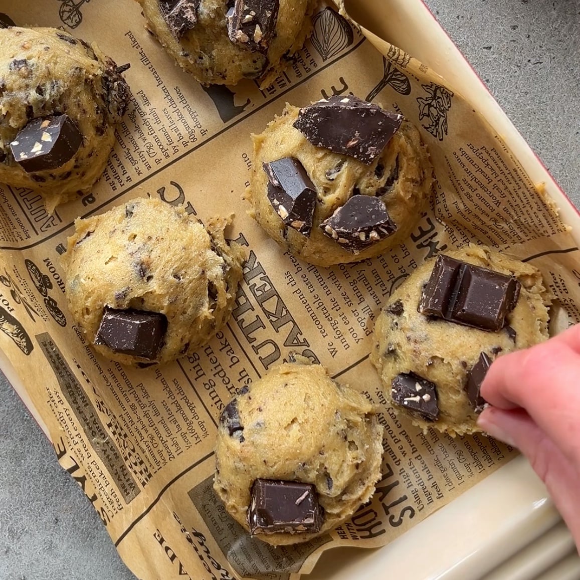 A hand is placing a chocolate chunk on a cookie dough scoop.