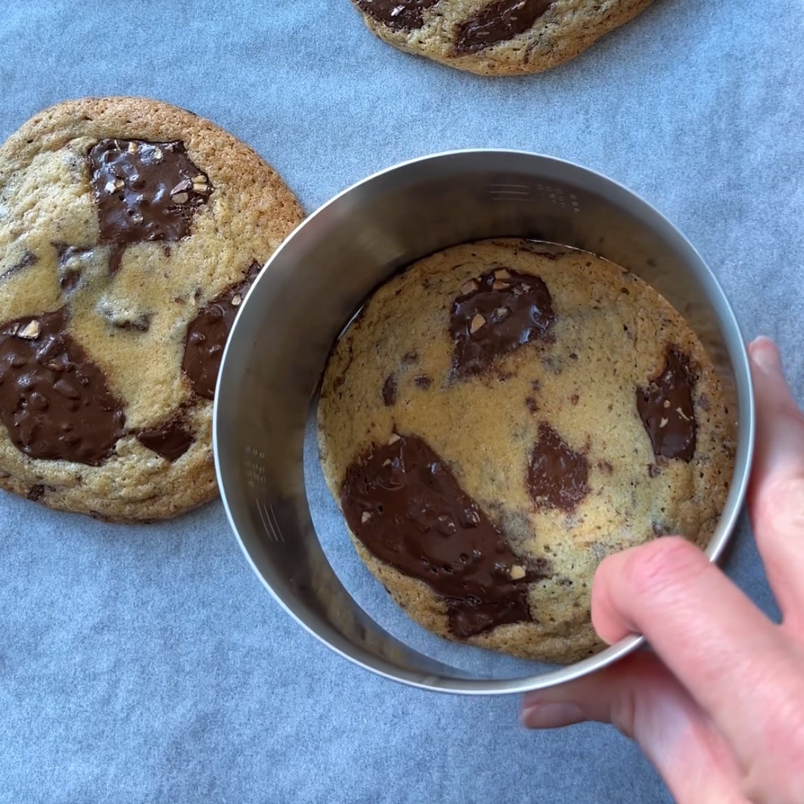 A hand uses a metal ring to shape a freshly baked chewy chocolate chip cookie on parchment paper, with another cookie nearby.