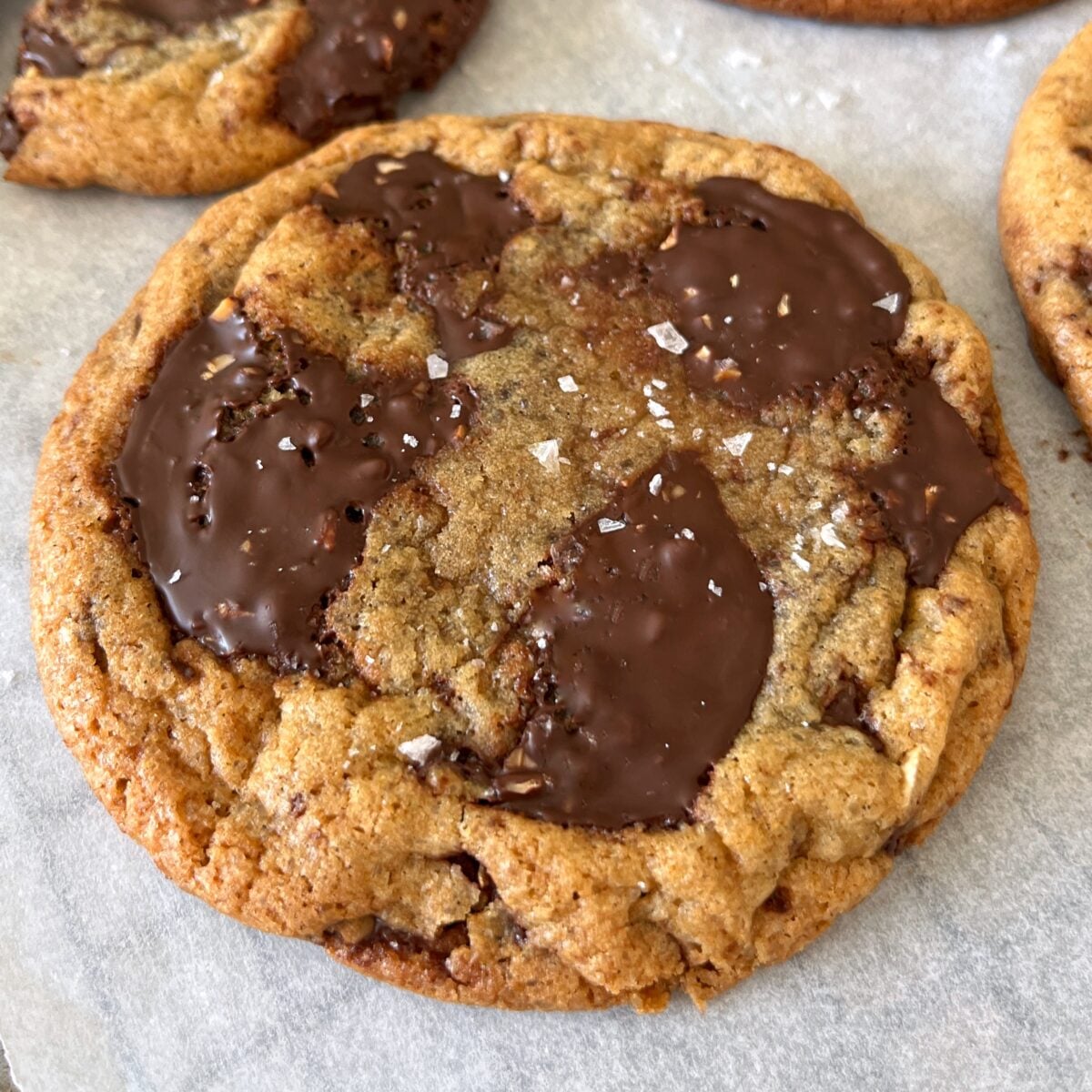 A chewy chocolate chip cookie with large melted chocolate chunks and a sprinkle of sea salt on top, resting on parchment paper.
