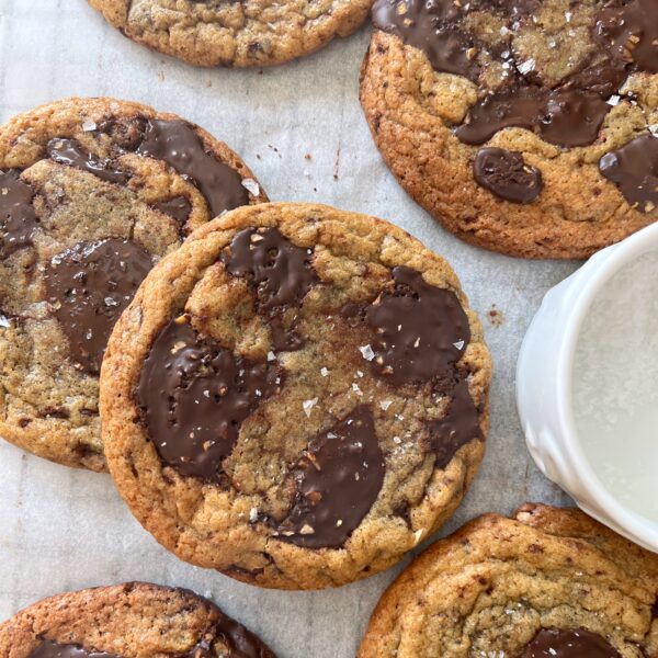 Chewy Chocolate Chip Cookies with large melted chocolate pieces and a sprinkling of sea salt are arranged on parchment paper next to a small white bowl of coarse salt.