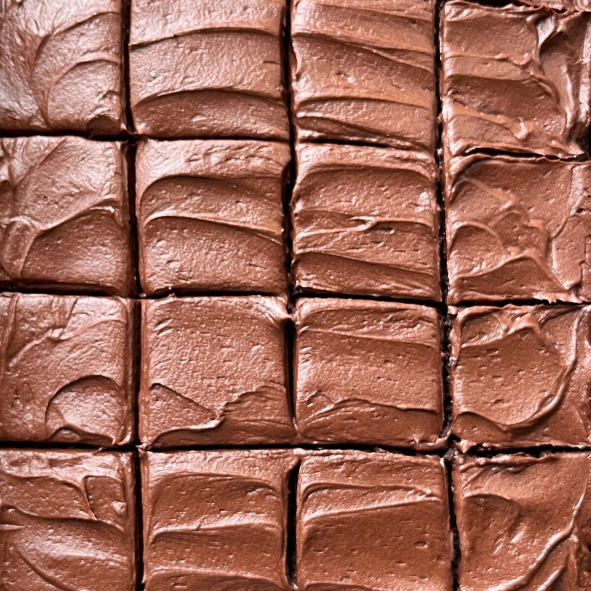 Rows of Chocolate Sheet Cake squares closely arranged, each piece topped with smooth, swirled chocolate icing.