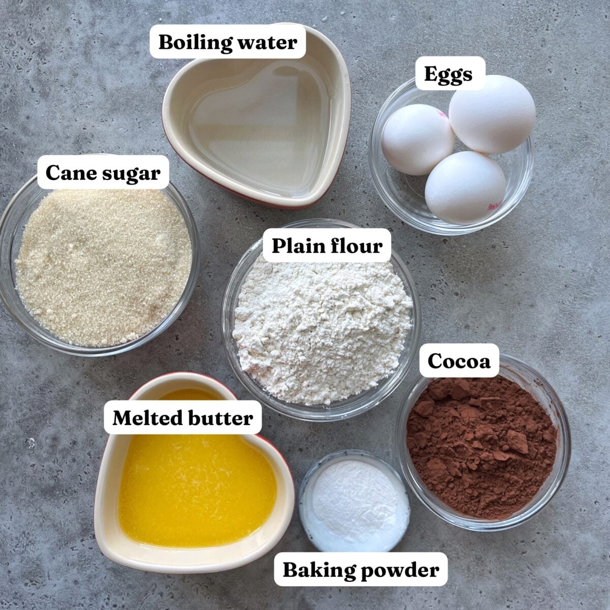 Bowls containing boiling water, eggs, cane sugar, plain flour, cocoa powder, melted butter, and baking powder are arranged on a gray surface with each ingredient labeled.