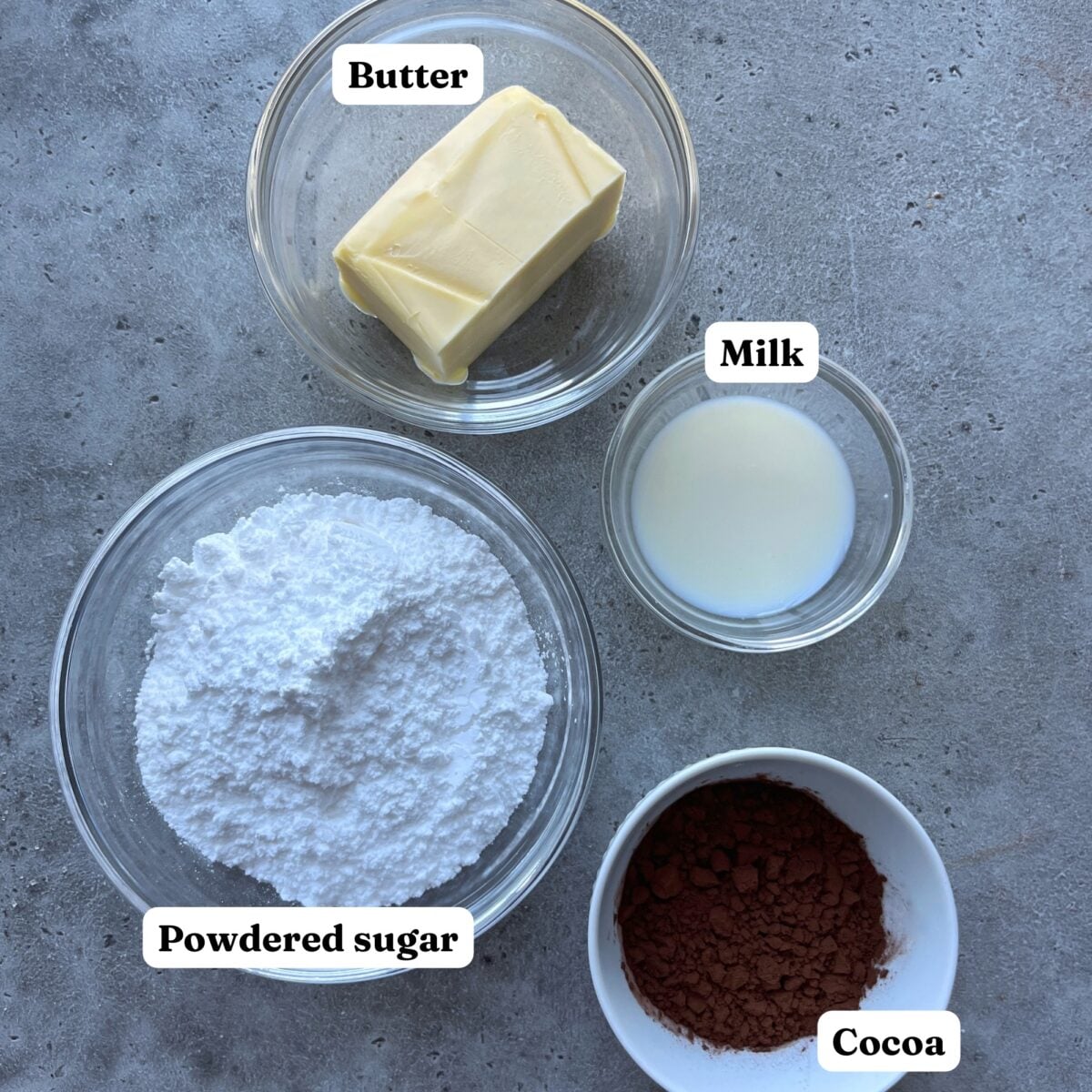 Four bowls with icing ingredients: butter, milk, powdered sugar, and cocoa powder. Each ingredient is labeled.