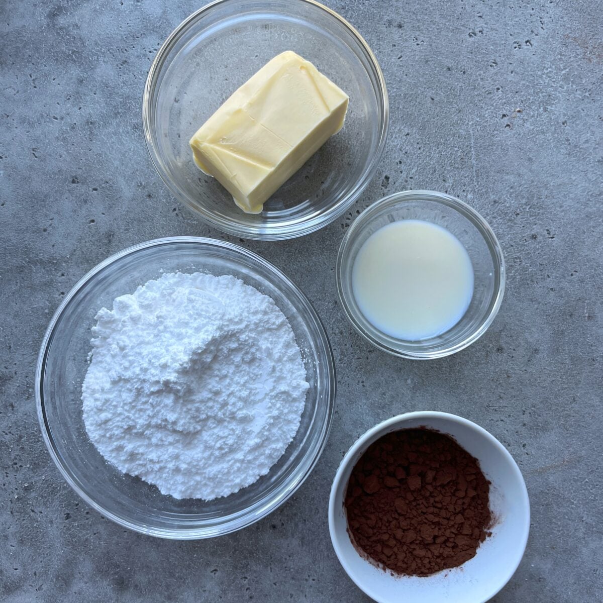 Four bowls with baking ingredients: butter, milk, powdered sugar, and cocoa powder.