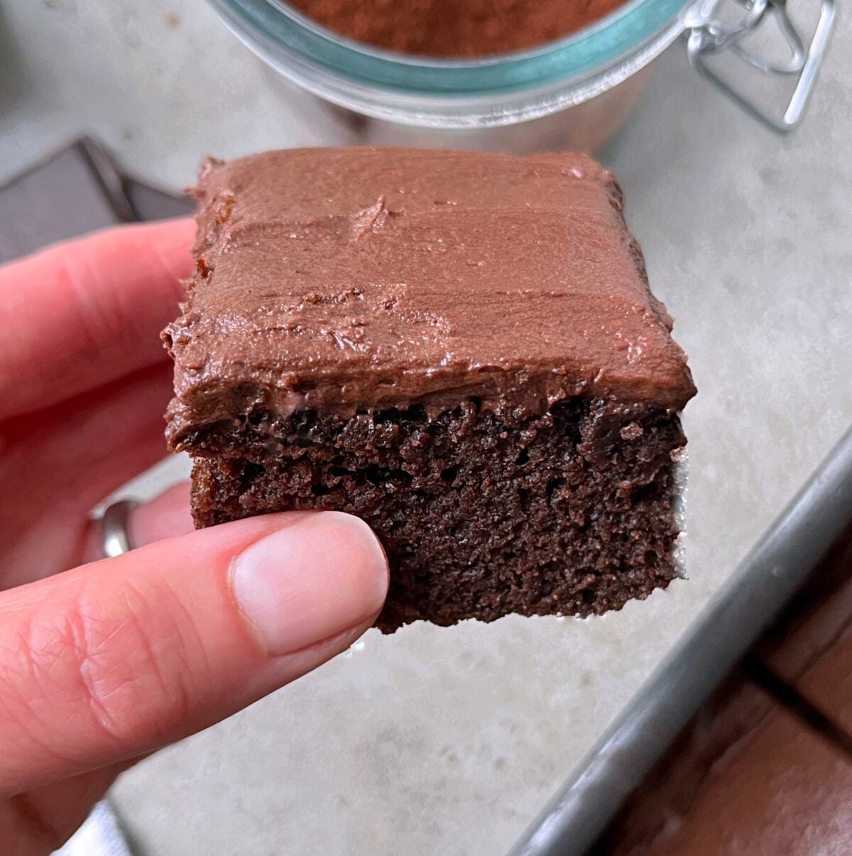 A hand holds a square piece of Chocolate Sheet Cake with chocolate frosting above a countertop.
