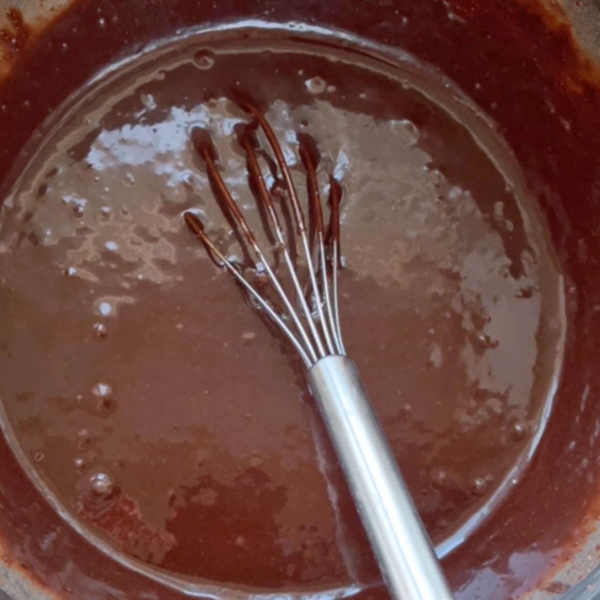 A metal whisk rests in a bowl of glossy chocolate batter, with small air bubbles visible on the surface.