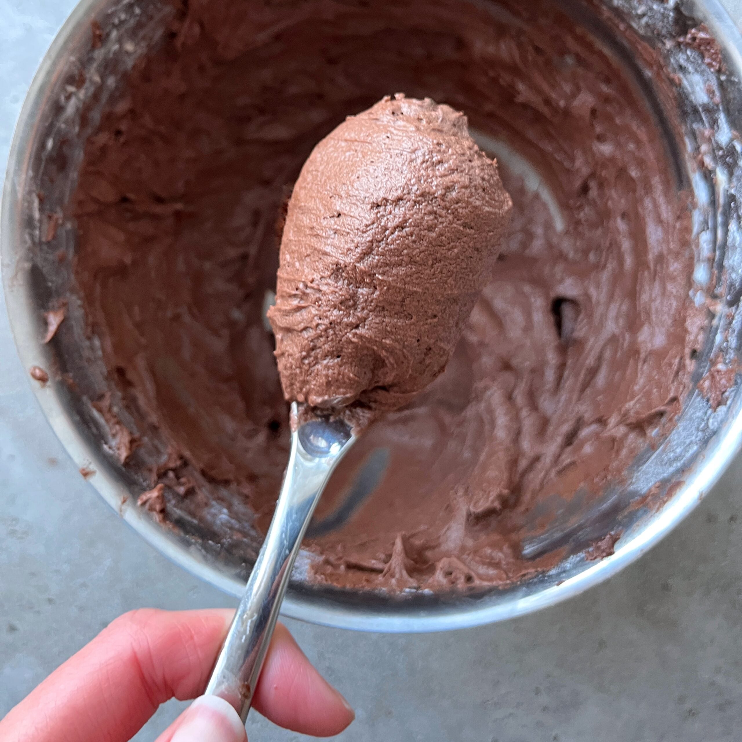 A hand holds a spoonful of chocolate icing above a metal bowl filled with more icing.