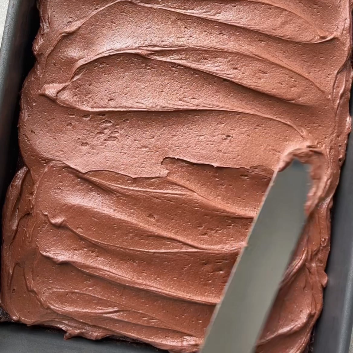 A palet knife spreads chocolate frosting evenly over a freshly baked Chocolate Sheet Cake.