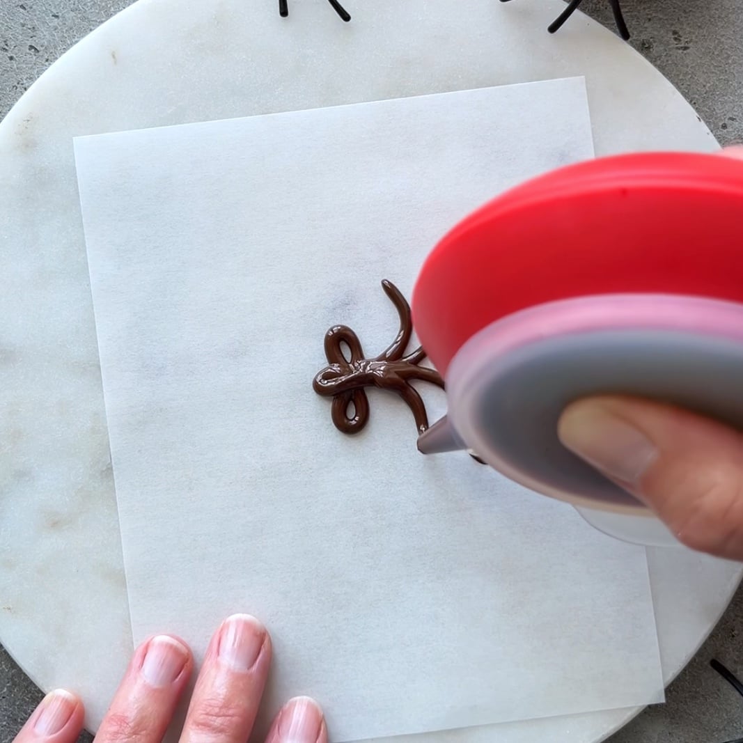 A hand uses a red piping tool to pipe chocolate onto parchment paper.